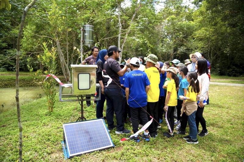 A large group of schoolchildren gather round water and rain level monitoring equipment on a riverbank as they listen to Hub researchers 